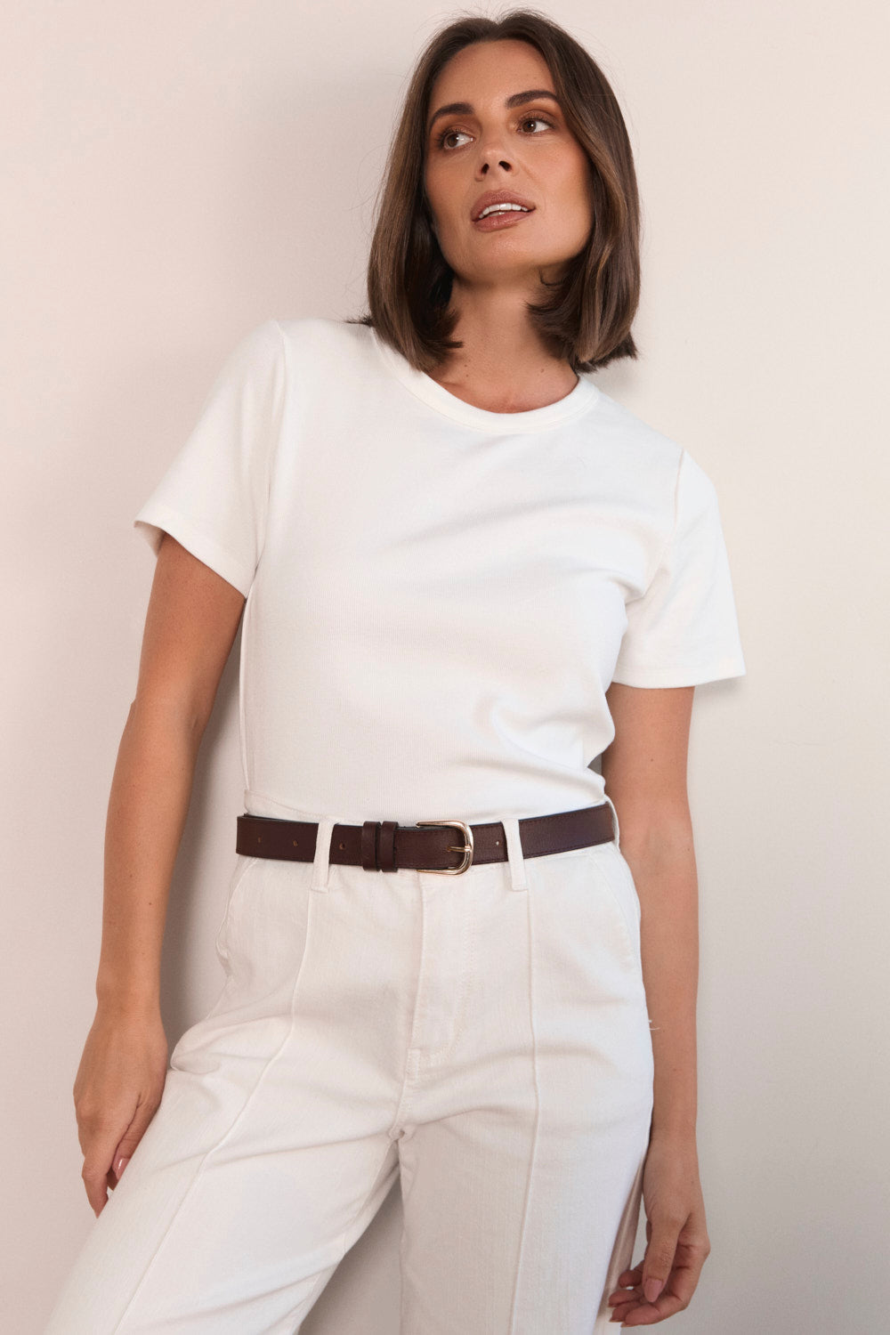 Woman wearing a white t-shirt and light-colored pants with a brown belt against a plain background