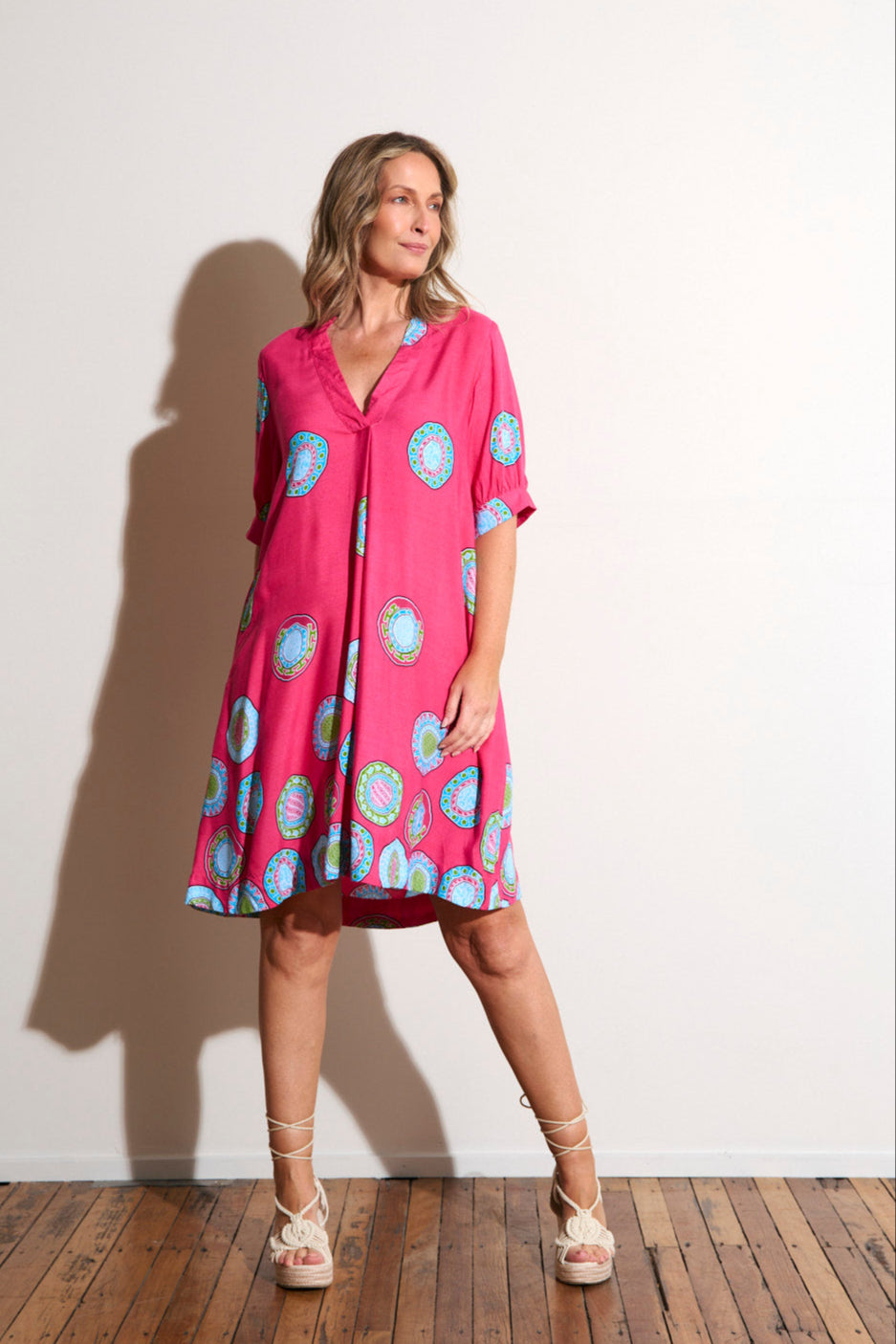 Woman wearing a pink dress with blue patterns against a white wall.