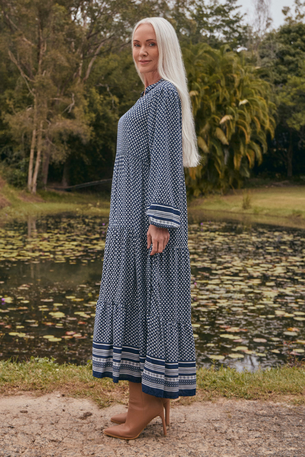 Woman in a blue patterned dress standing by a pond with greenery in the background