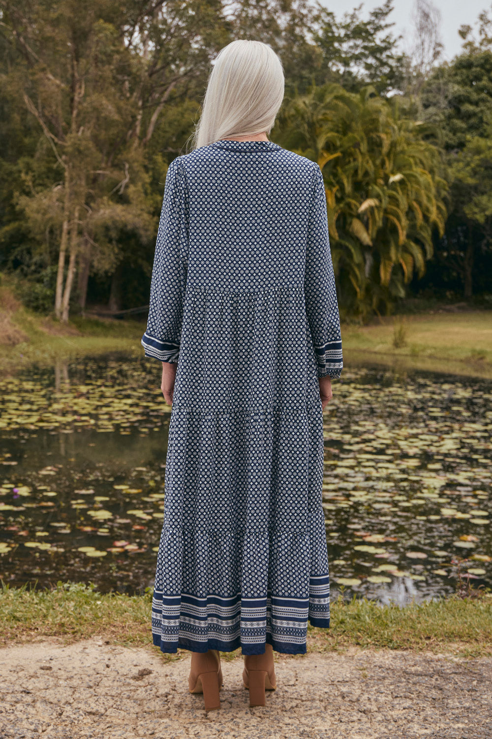Person wearing a long blue dress with white patterns standing by a pond.