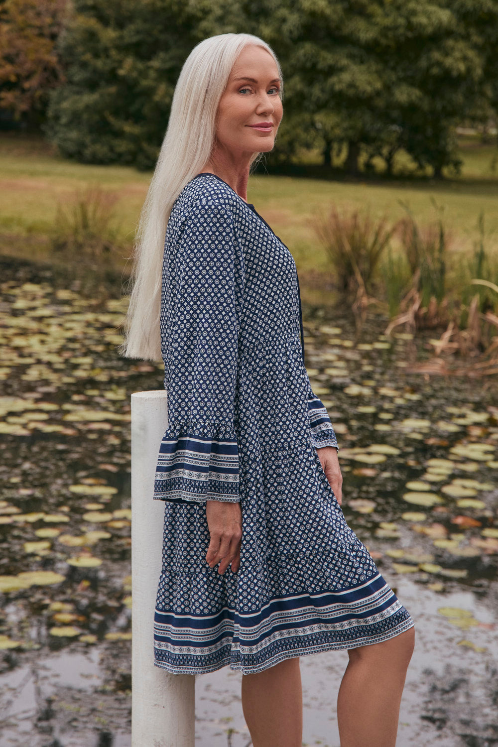 Woman in a blue patterned dress standing by a pond with trees in the background