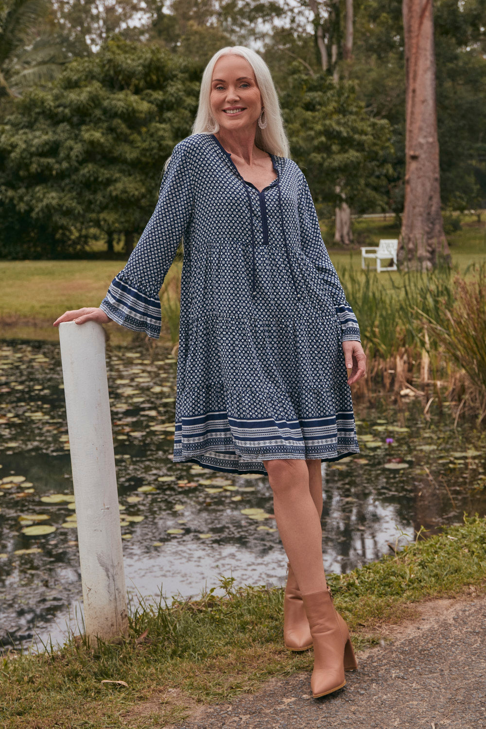 Woman in a blue patterned dress standing by a pond with trees in the background