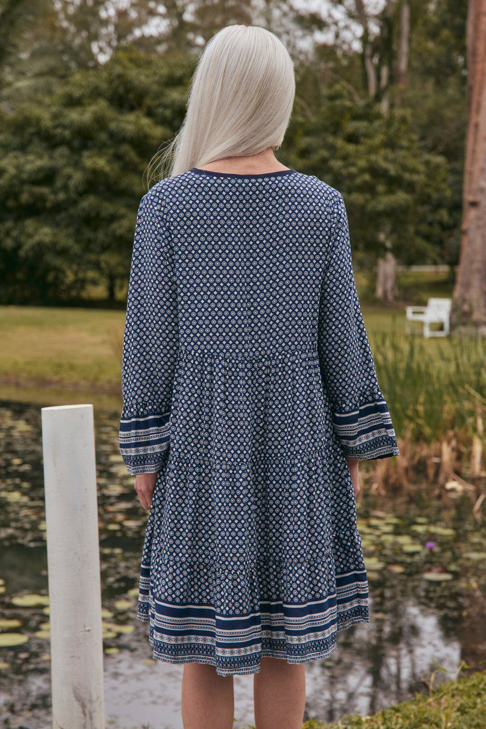 Person wearing a blue patterned dress standing by a pond with trees in the background