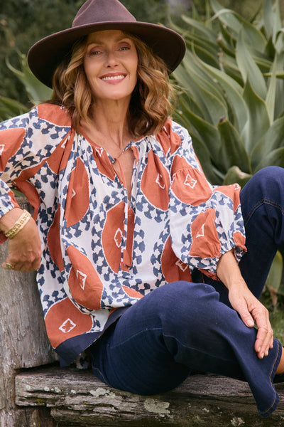 Woman wearing a patterned blouse and wide-brimmed hat sitting outdoors.