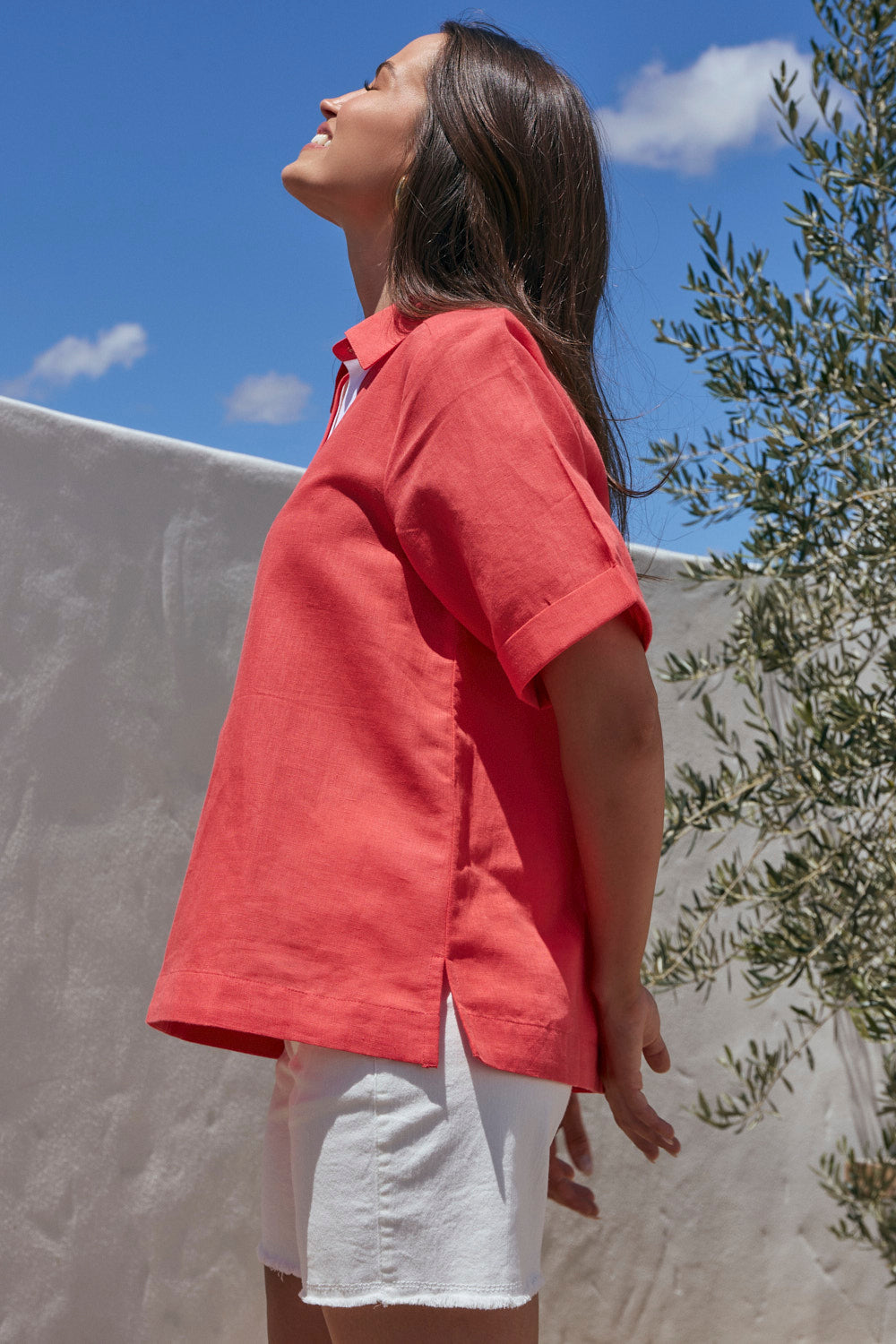 Woman in a red shirt and white shorts standing outdoors with a clear blue sky and olive tree in the background.