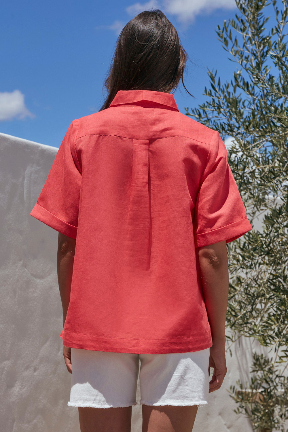 Person wearing a red shirt and white shorts standing outdoors with a blue sky and greenery in the background.