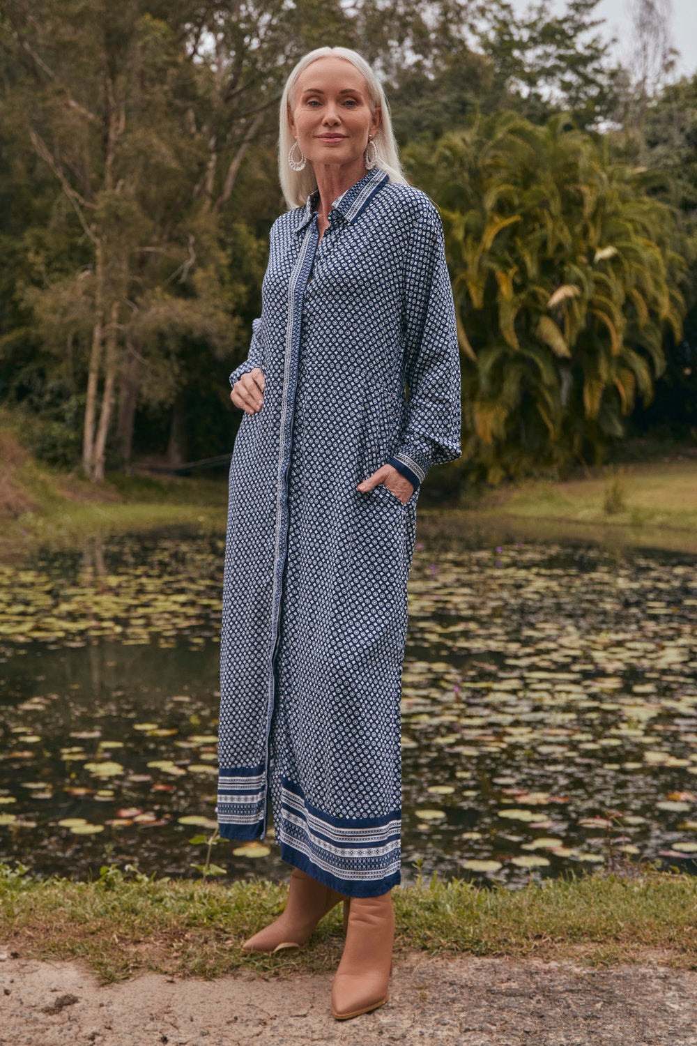 Woman in a blue patterned dress standing by a pond with greenery in the background