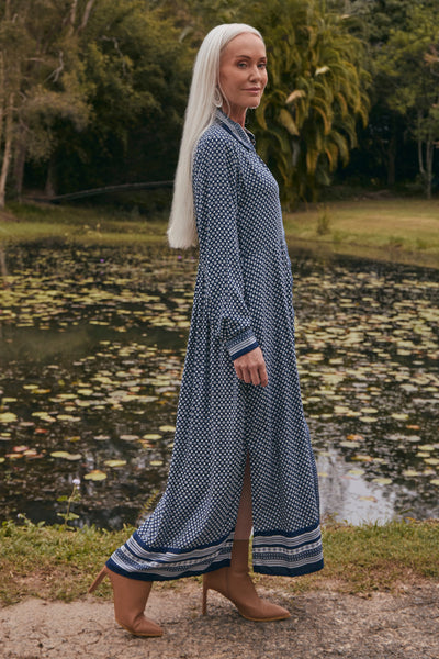 Woman in a long blue patterned dress standing by a pond with trees in the background