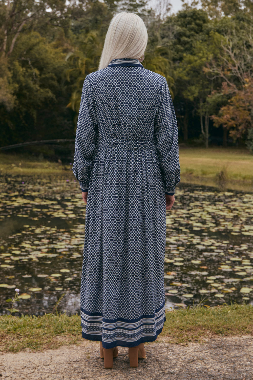 Person wearing a long, patterned dress standing by a pond with trees in the background