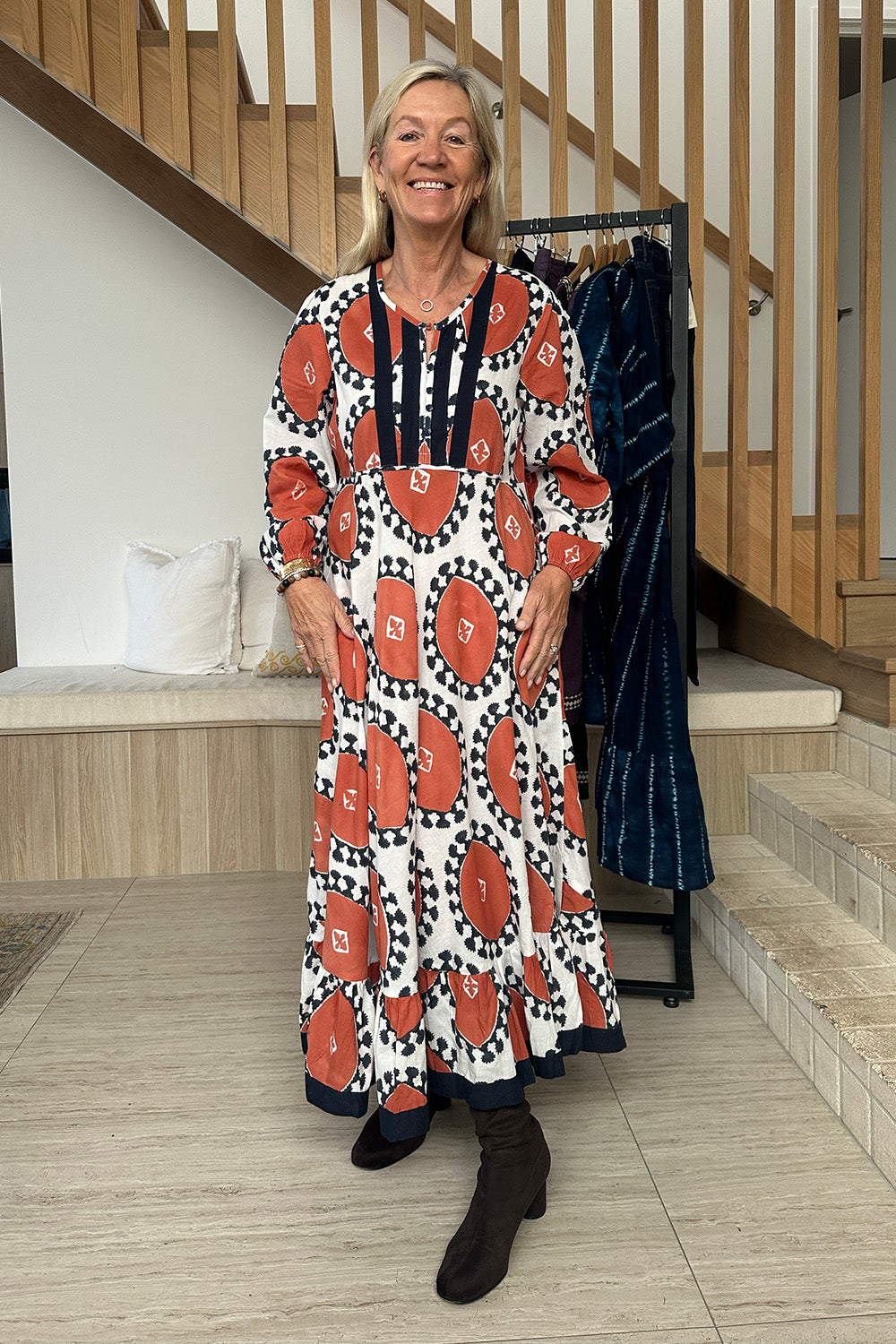 Woman in a patterned dress standing in a home setting with wooden stairs and a coat rack.