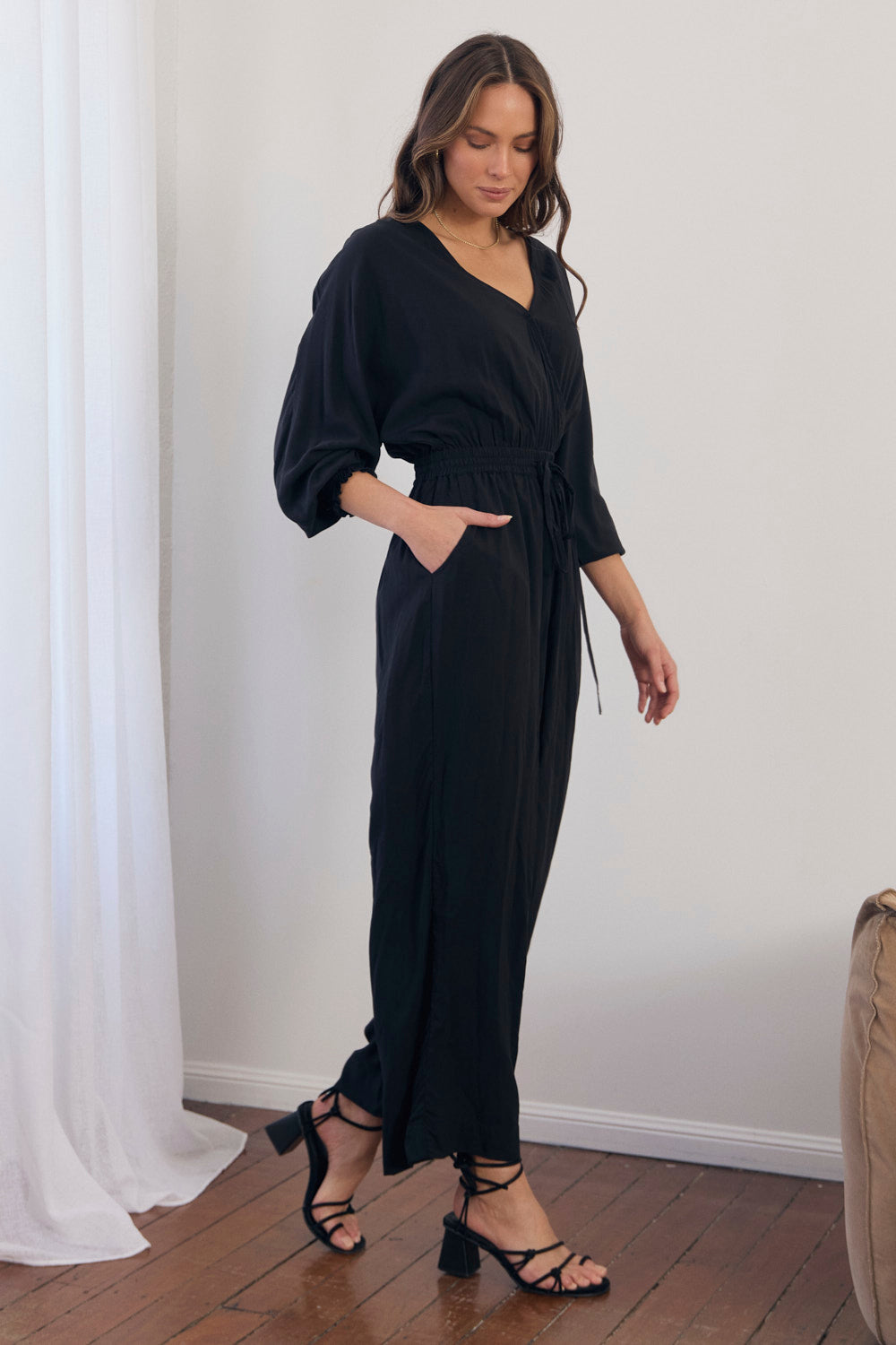 Woman wearing a black dress standing in a room with white curtains and wooden floor.