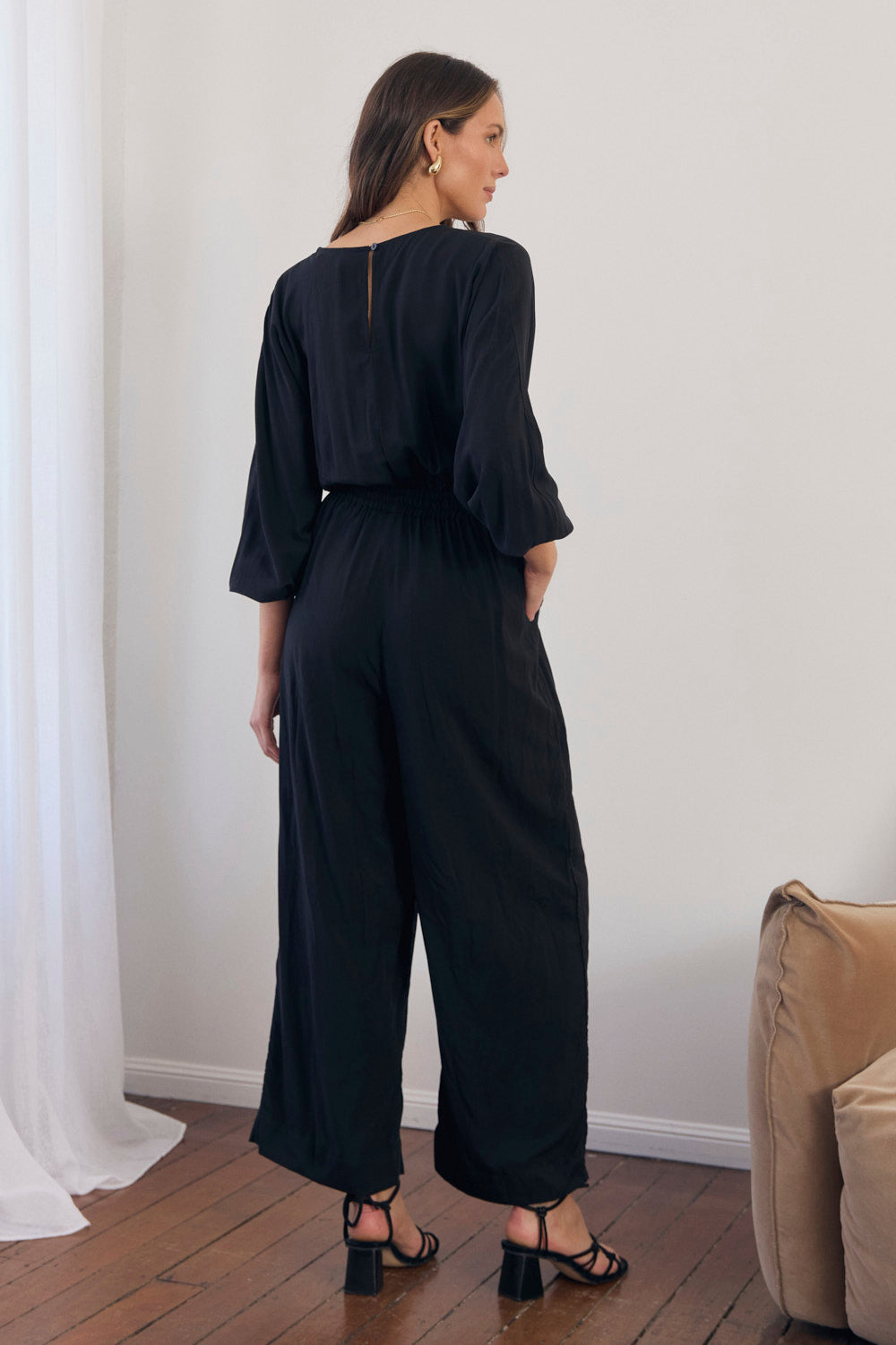 Woman wearing a black jumpsuit standing in a room with white walls and a beige couch.